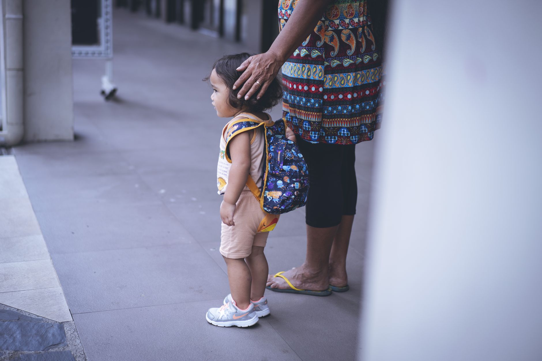 Un jeune enfant avec un sac à dos se tient à côté de la touche protectrice d'un soignant à l'extérieur.