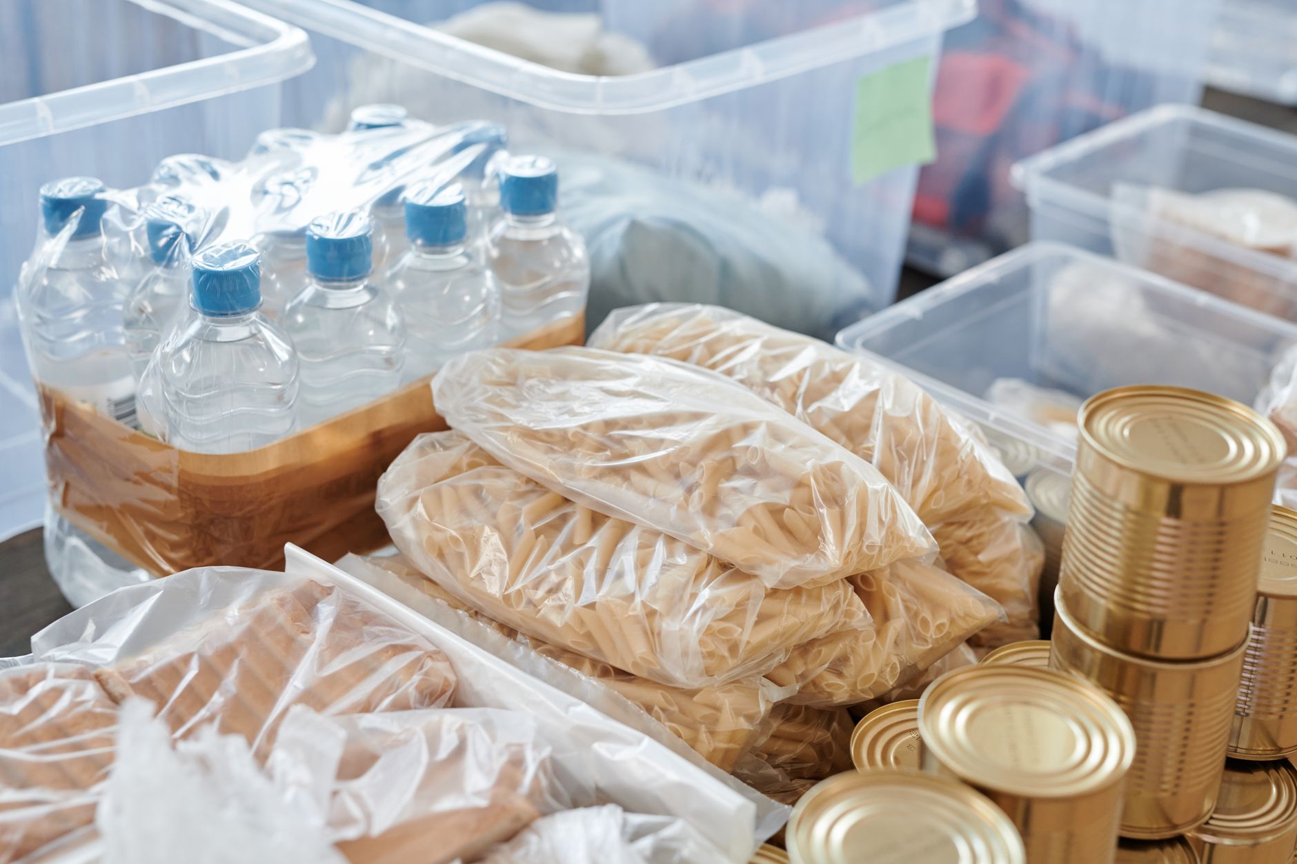 Sachets de pâtes, bouteilles d’eau sous film plastique et boîtes de conserve rangés dans des bacs en plastique transparents.