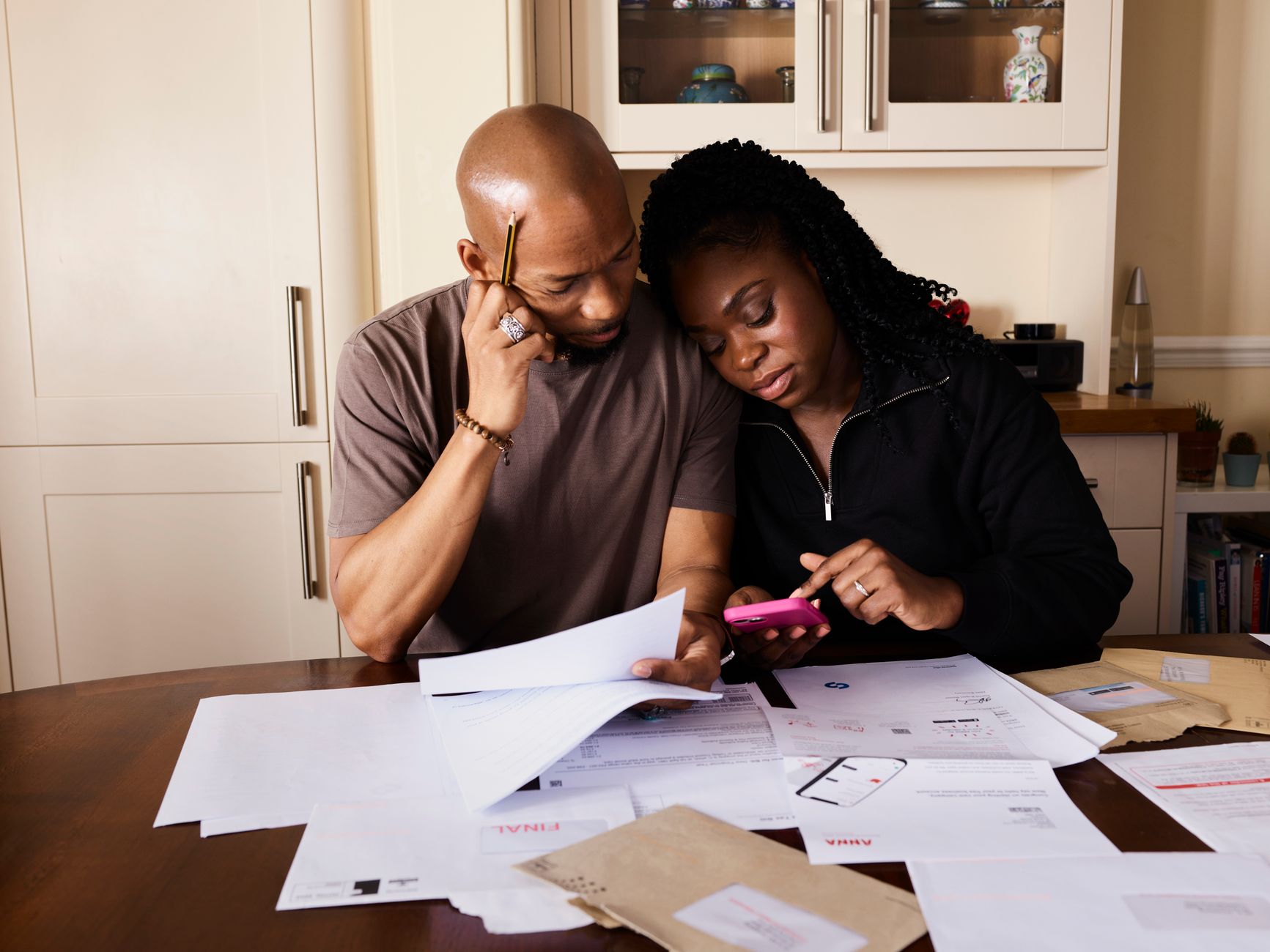 Un couple assis à une table examine des documents papier et des enveloppes, l’un tenant une feuille, l’autre un téléphone rose.