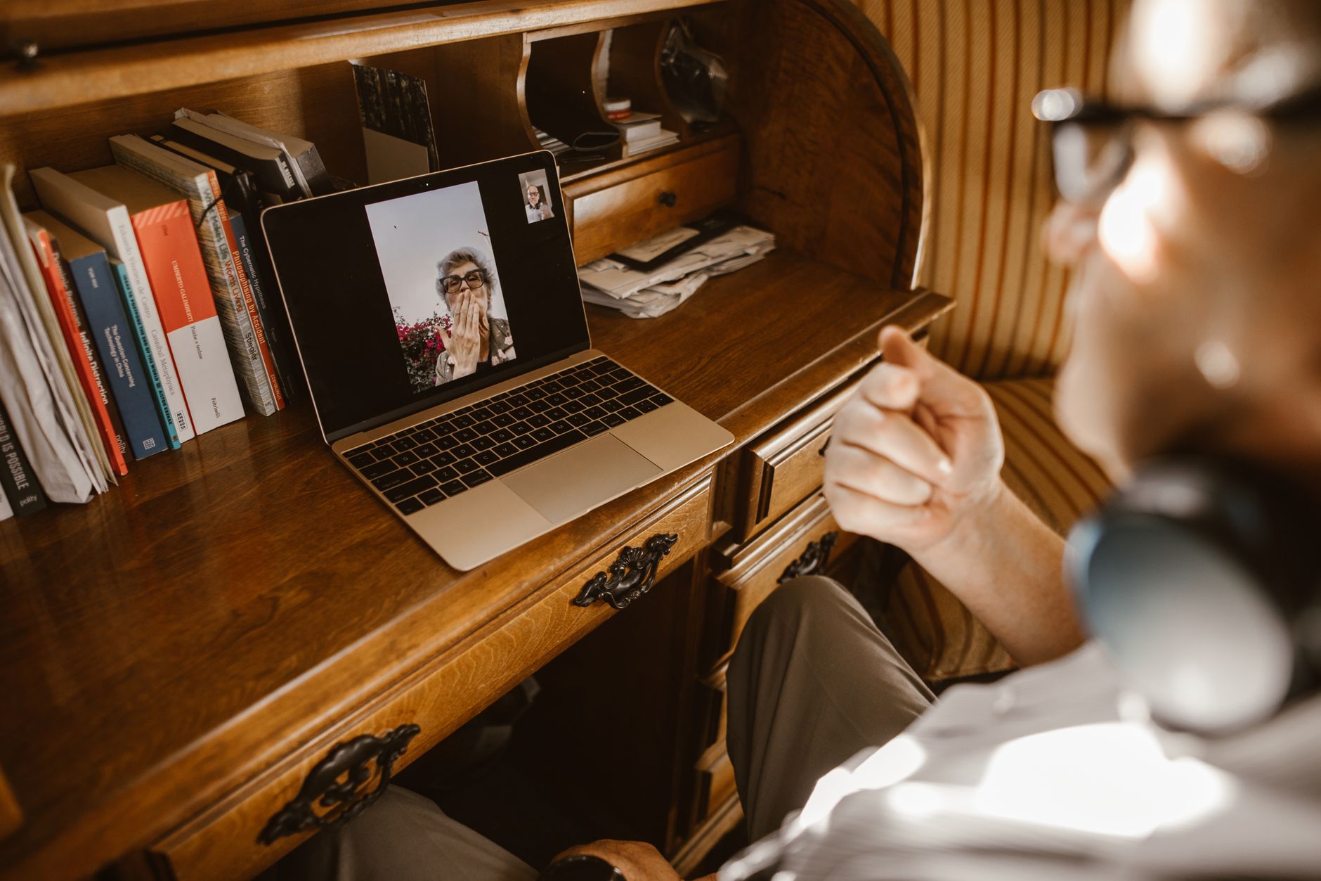 Un homme assis à un bureau en bois suit un appel vidéo sur un ordinateur portable où apparaît une femme âgée à lunettes.