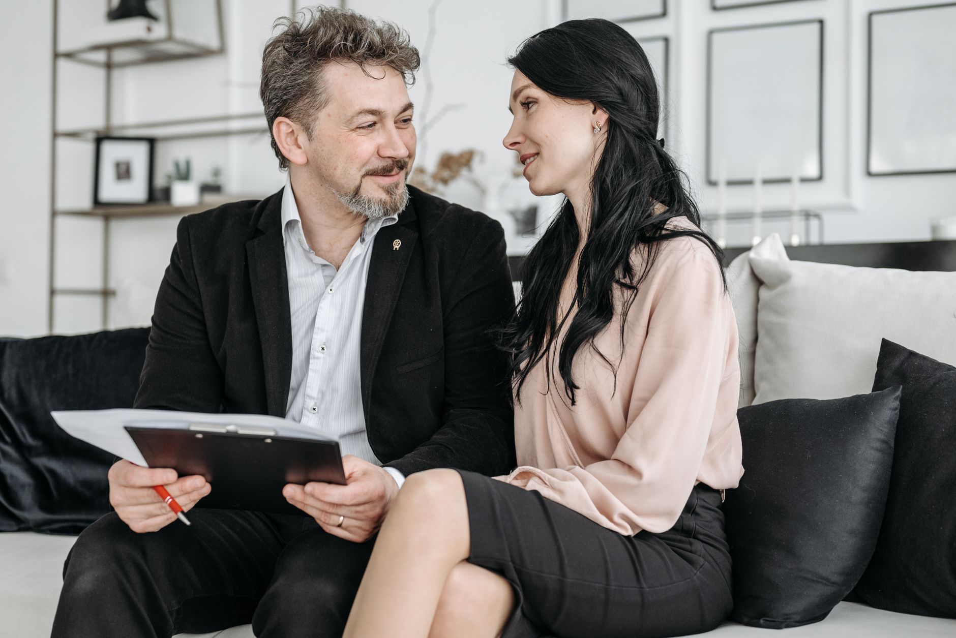 Un homme et une femme assis sur un canapé se sourient, l’homme tenant un presse-papiers avec des documents et un stylo rouge.