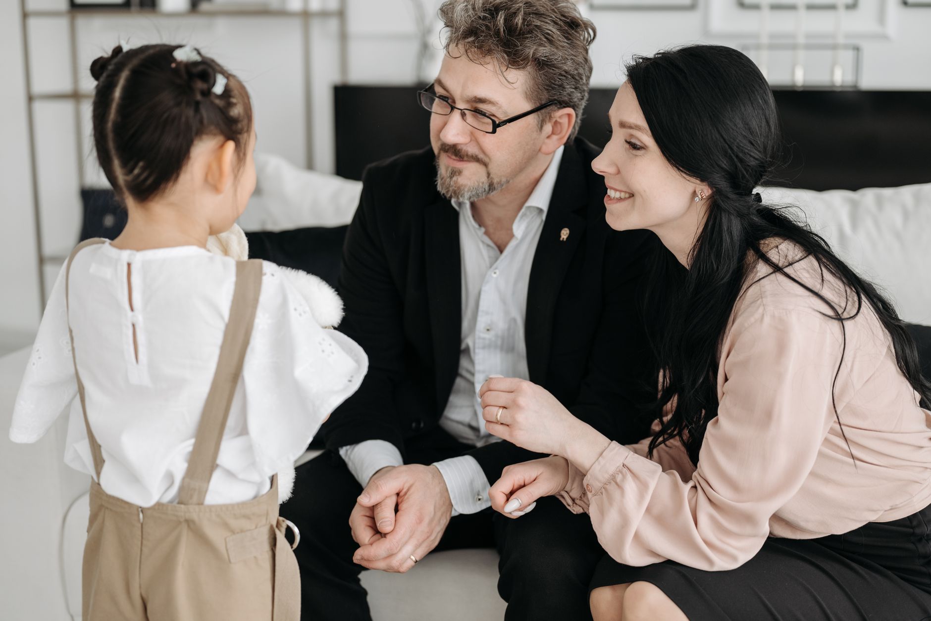 Un homme et une femme souriants, assis sur un canapé, parlent à une petite fille vue de dos dans un intérieur clair.