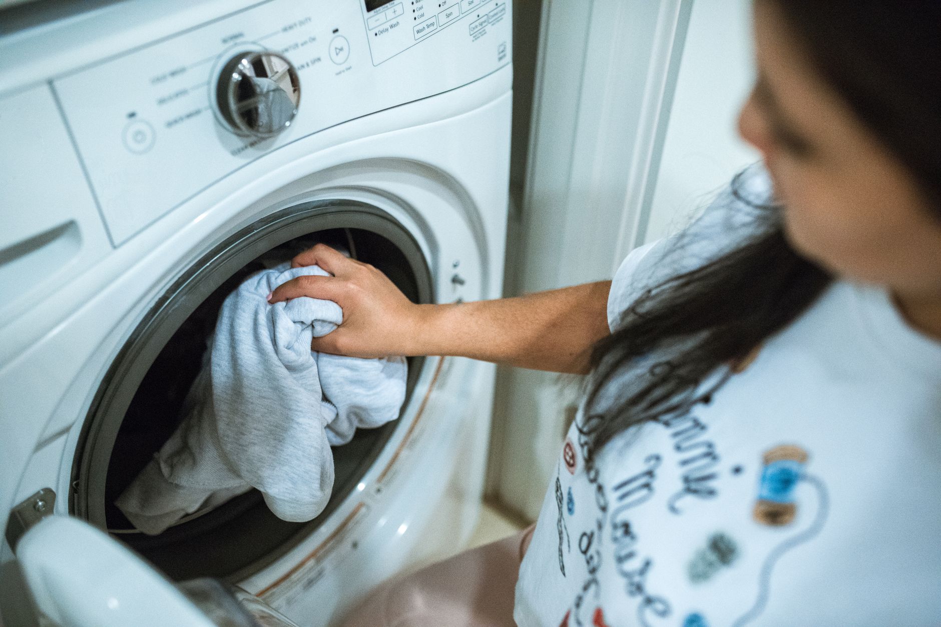 Une personne aux cheveux longs met du linge gris clair dans le hublot d’un lave-linge blanc à chargement frontal, dans un intérieur domestique.